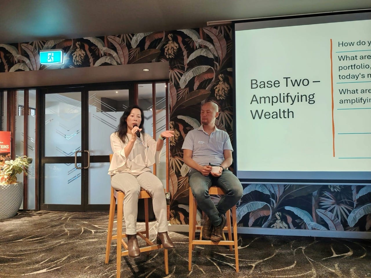 Two panelists on stools beside a slide titled Base Two, Amplifying Wealth